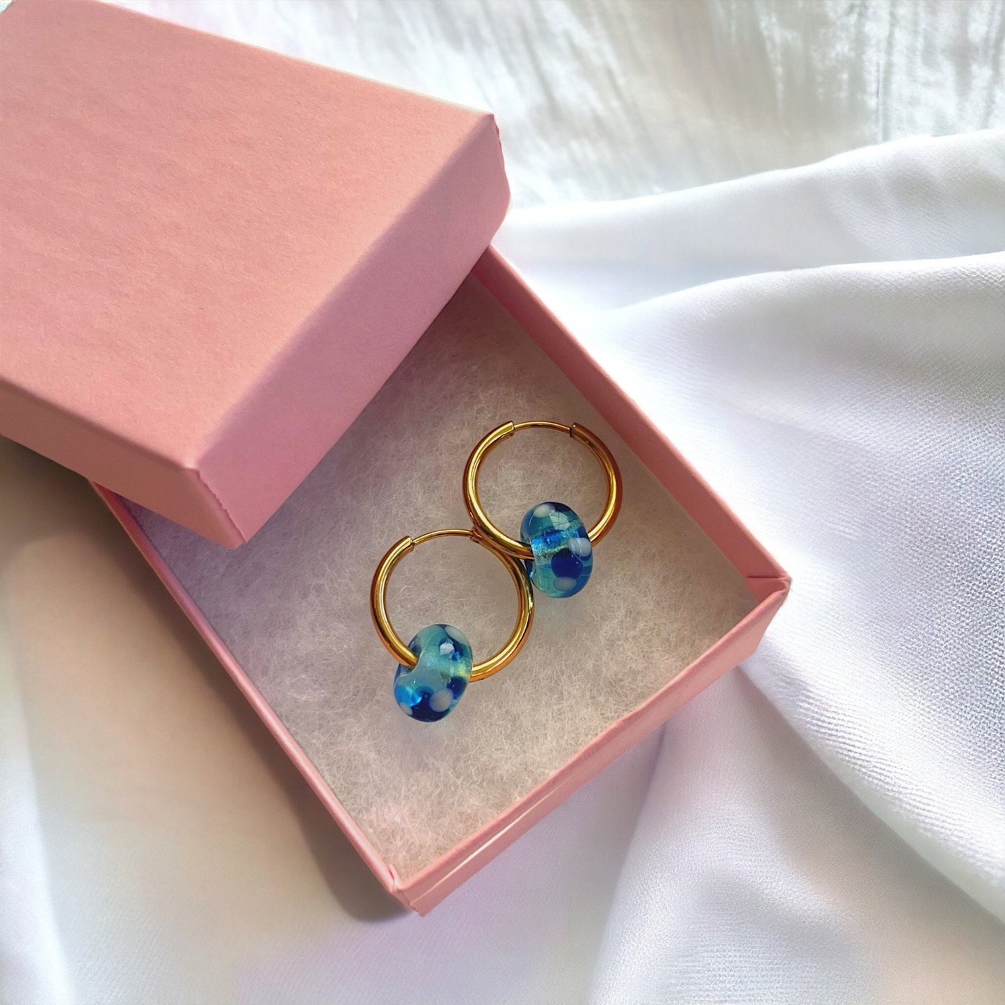 
                  
                    Gold hoop earrings with blue beads in a pink jewelry box on a white fabric background
                  
                