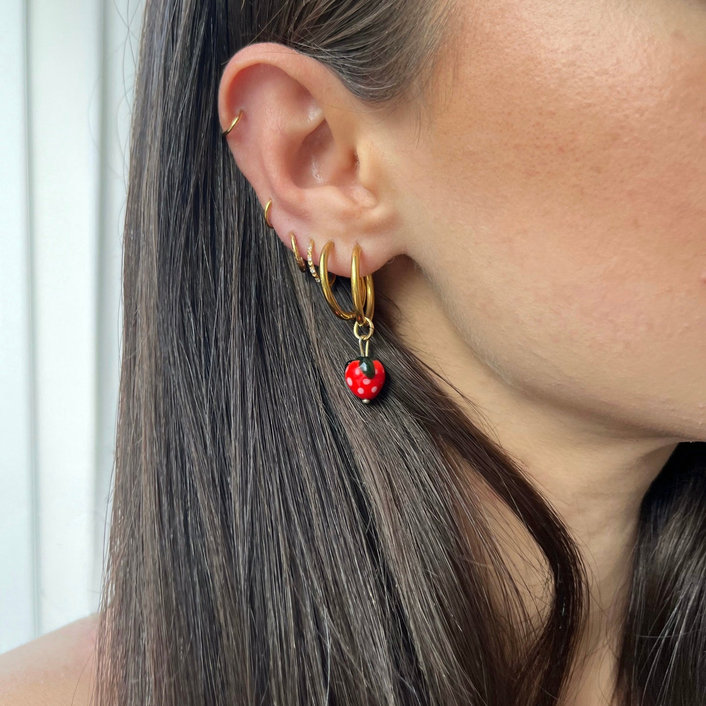 
                  
                    Close-up of an ear with gold hoop earrings featuring red heart charms.
                  
                