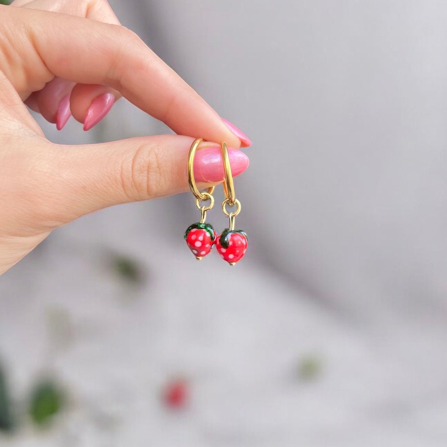 
                  
                    Hand holding gold earrings with strawberry-shaped charms against a blurred background
                  
                