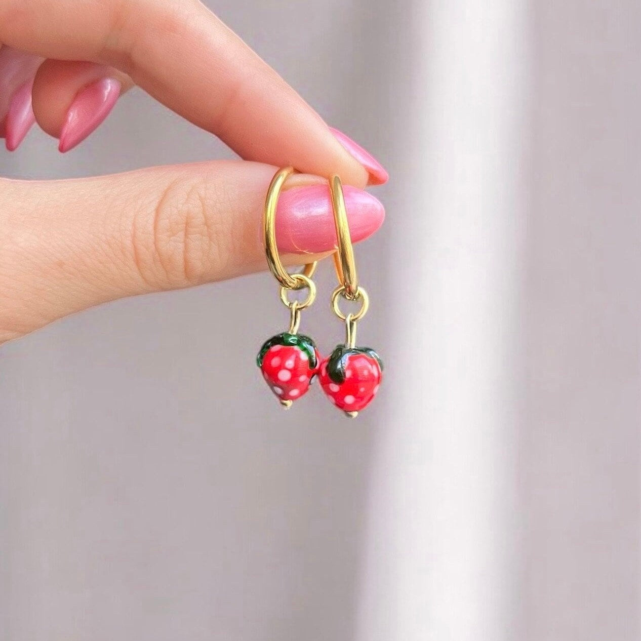 
                  
                    Gold hoop earrings with red strawberry charms held by a hand against a neutral background
                  
                