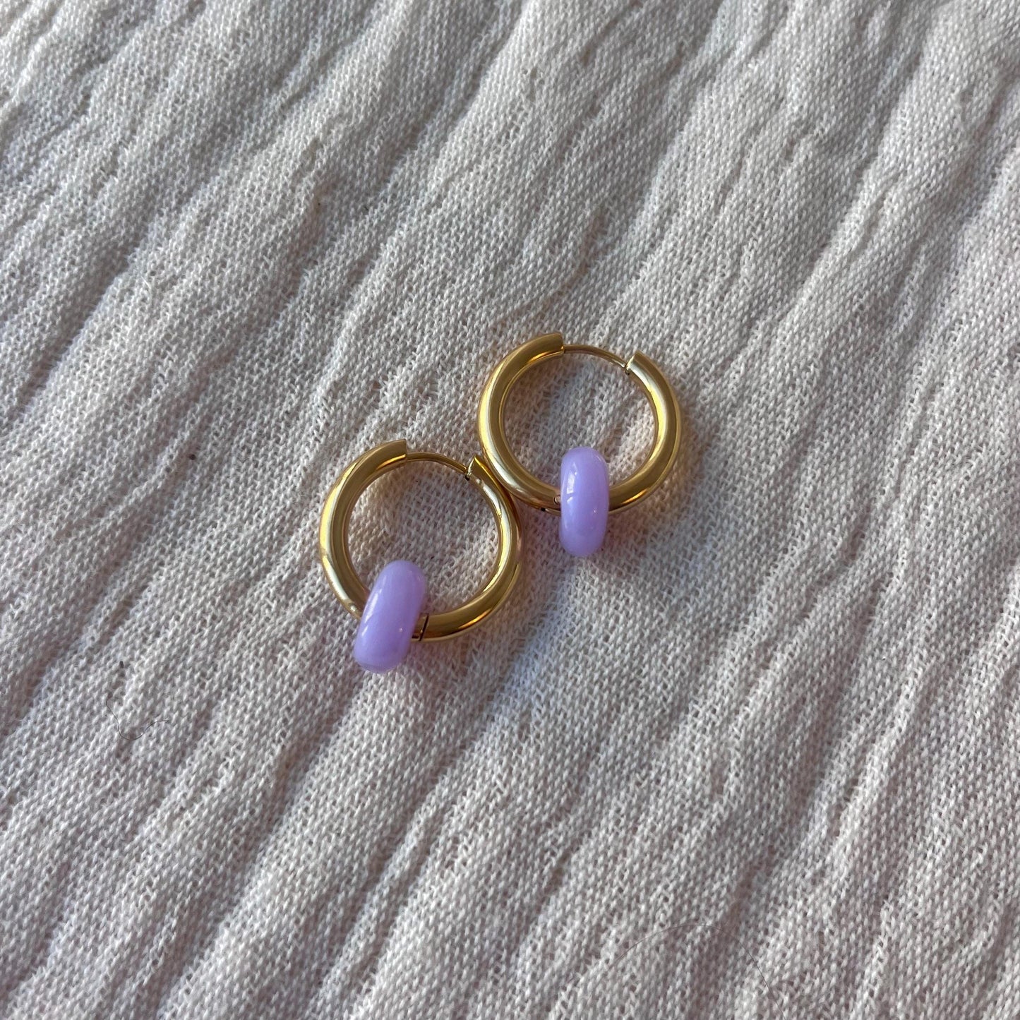 
                  
                    Gold hoop earrings with purple beads placed on fabric background
                  
                