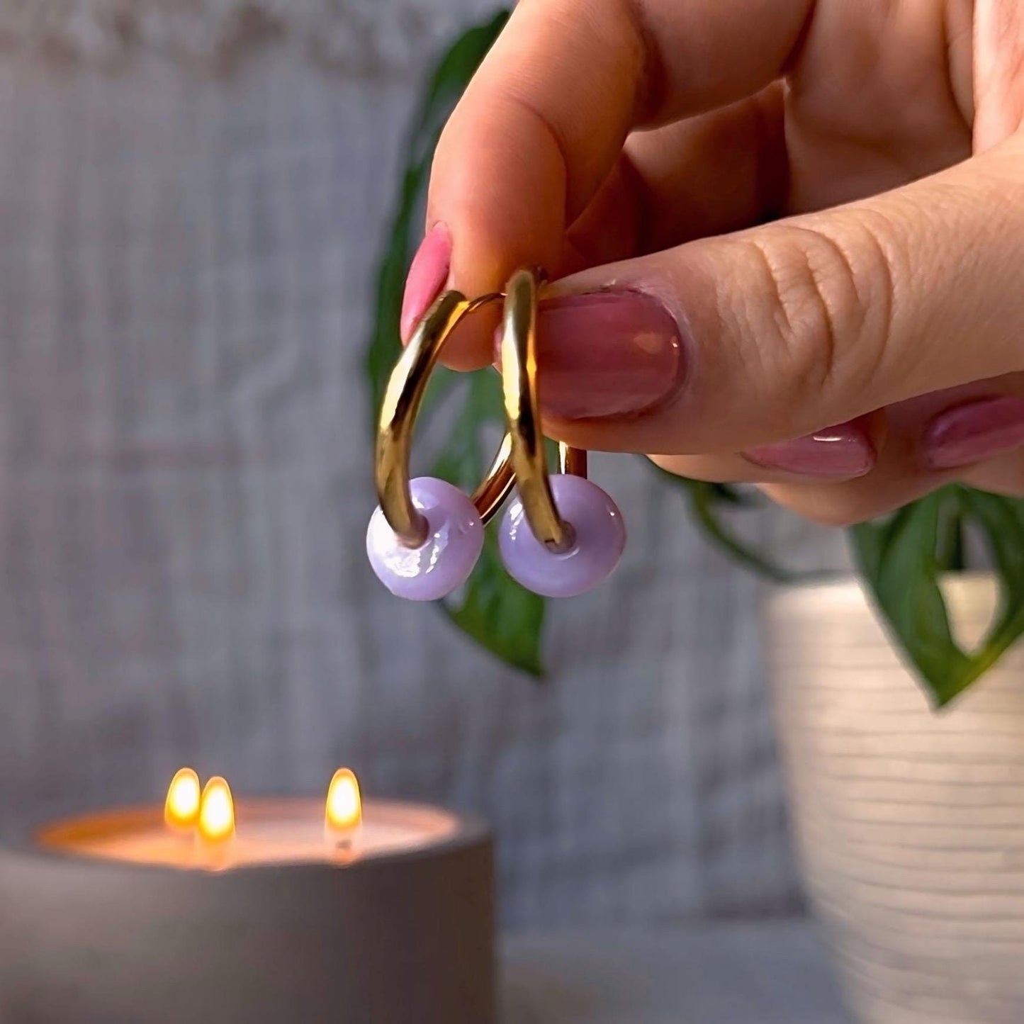Gold hoop earrings with purple beads held by a hand in front of a candle.