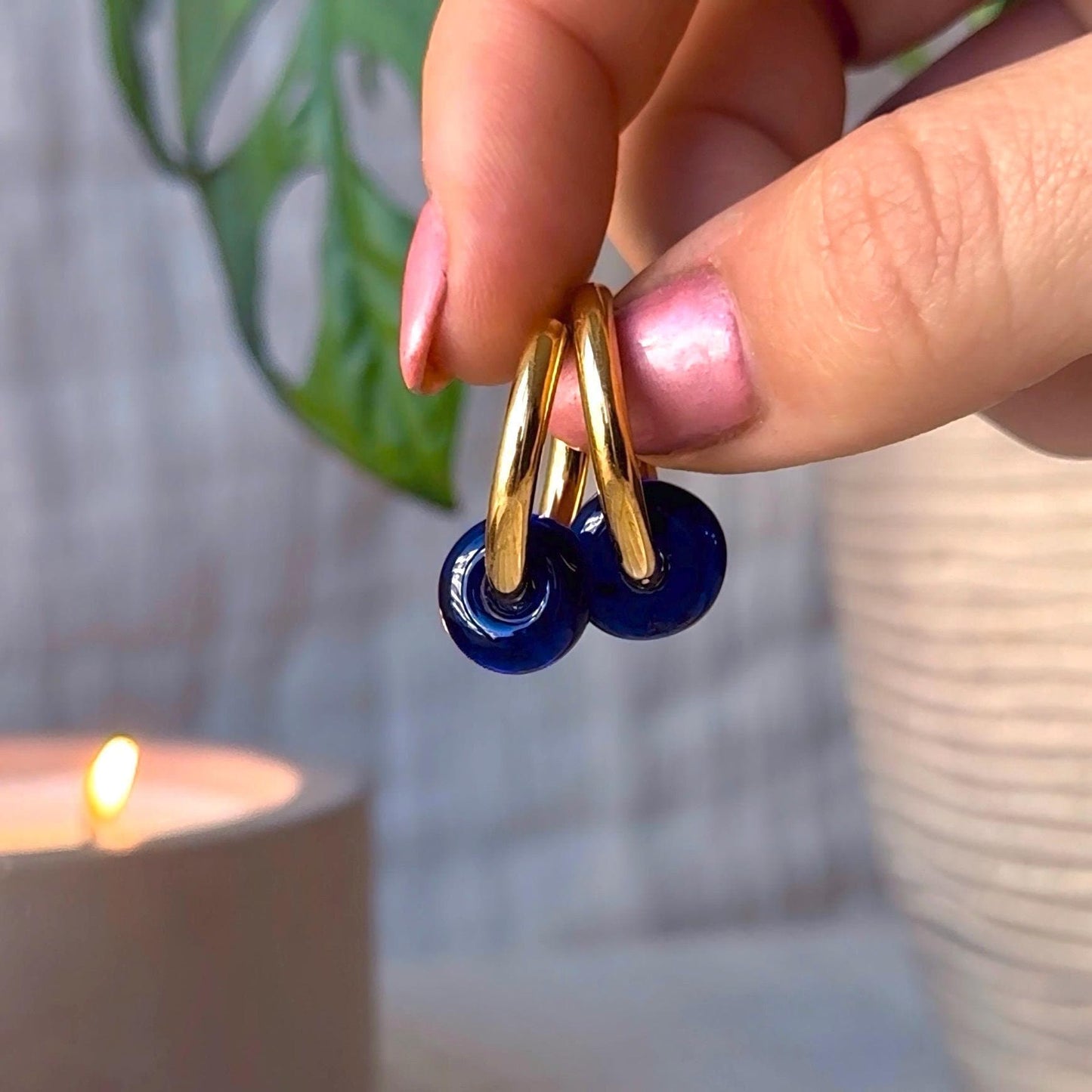 Gold and blue earrings held by a hand with pink nail polish, blurred background