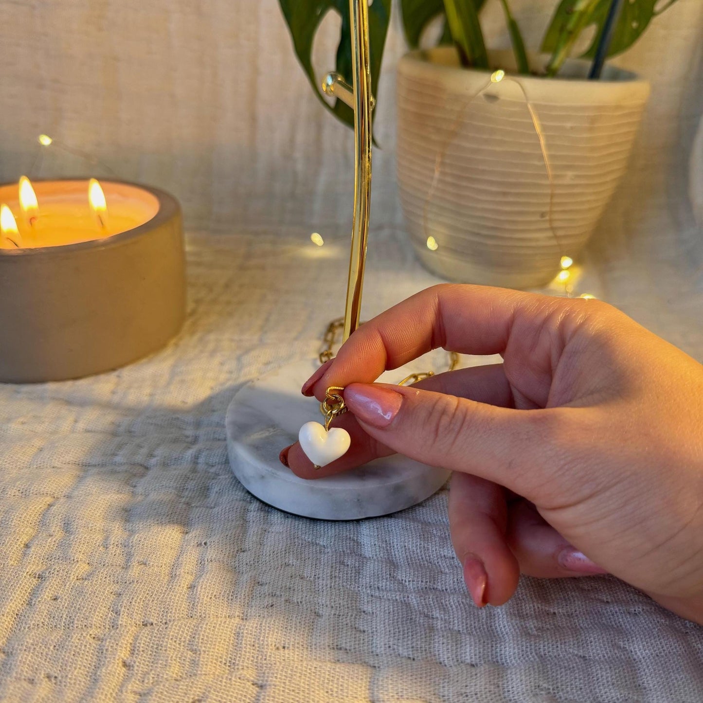 
                  
                    Hand holding a heart-shaped pendant with a candle and plant in the background
                  
                