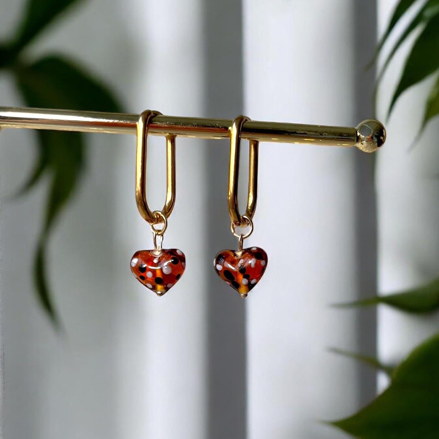 Gold hoop earrings with tortoiseshell heart charms on a gold rod against a white background with green leaves.
