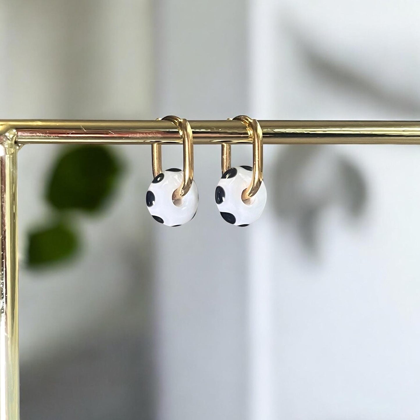 
                  
                    Pair of earrings with black and white polka dot design hanging on a gold rod against a neutral background.
                  
                