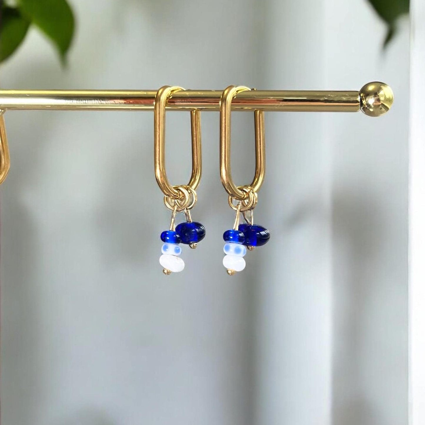 Gold hoop earrings with blue and white bead drops on a gold bar against a blurred natural background.