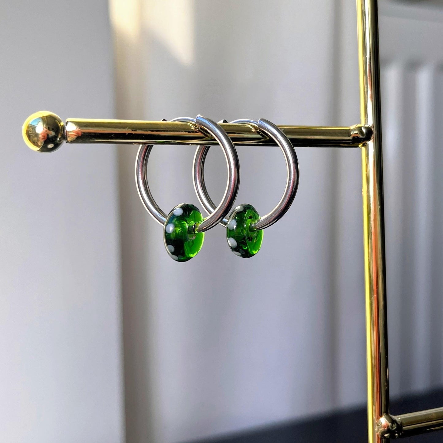 
                  
                    Silver hoop earrings with green gemstones on a gold stand against a white background
                  
                