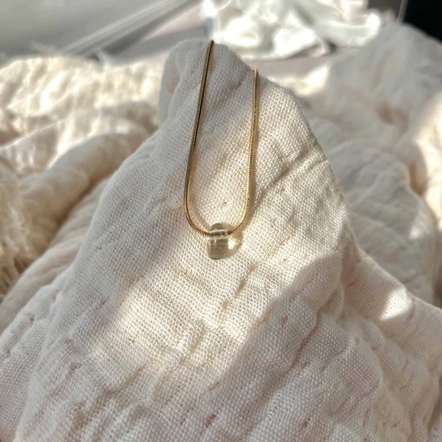 
                  
                    Gold necklace with a clear pendant on a textured white fabric background
                  
                