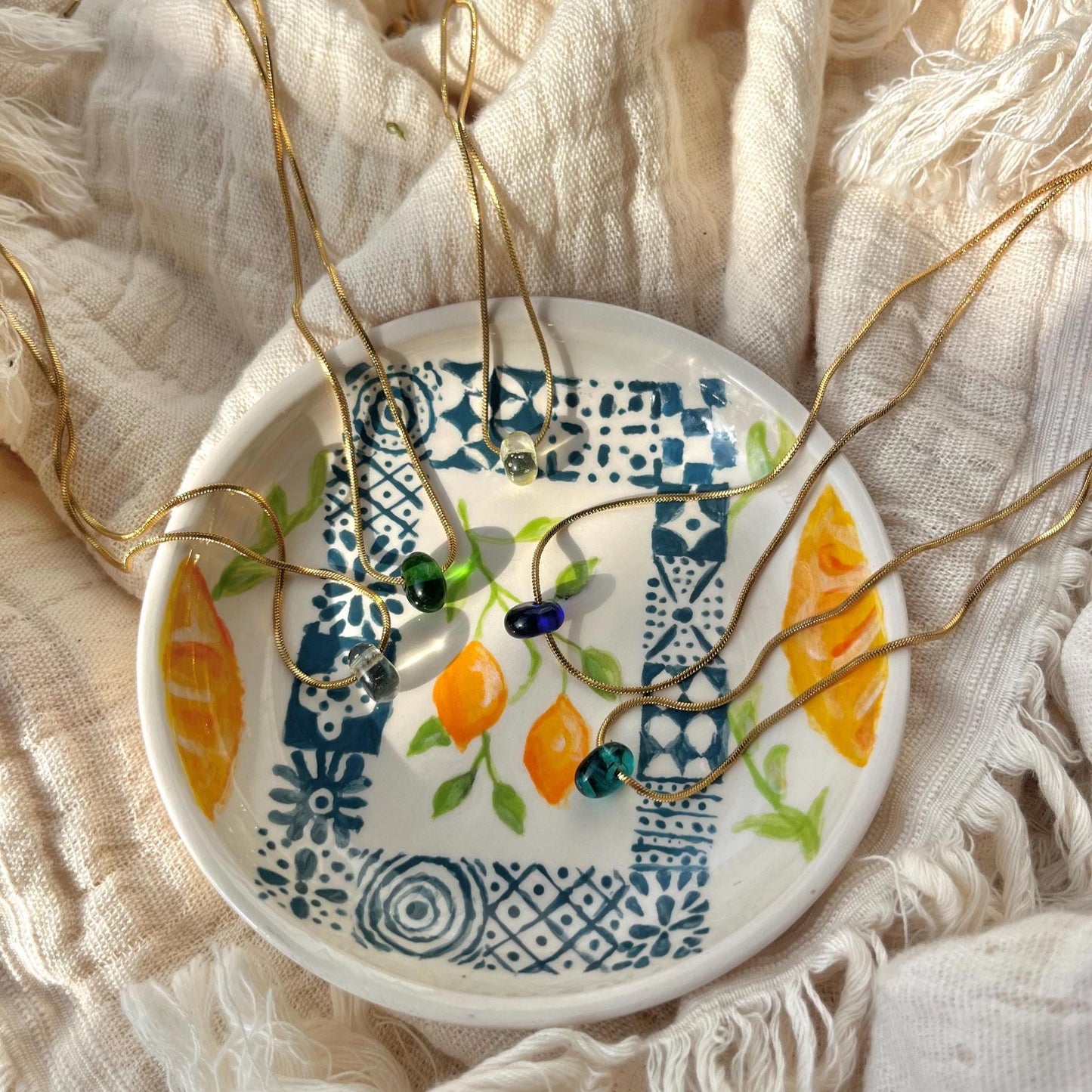 
                  
                    Decorative ceramic dish with floral patterns holding gold necklaces on a textured fabric background
                  
                