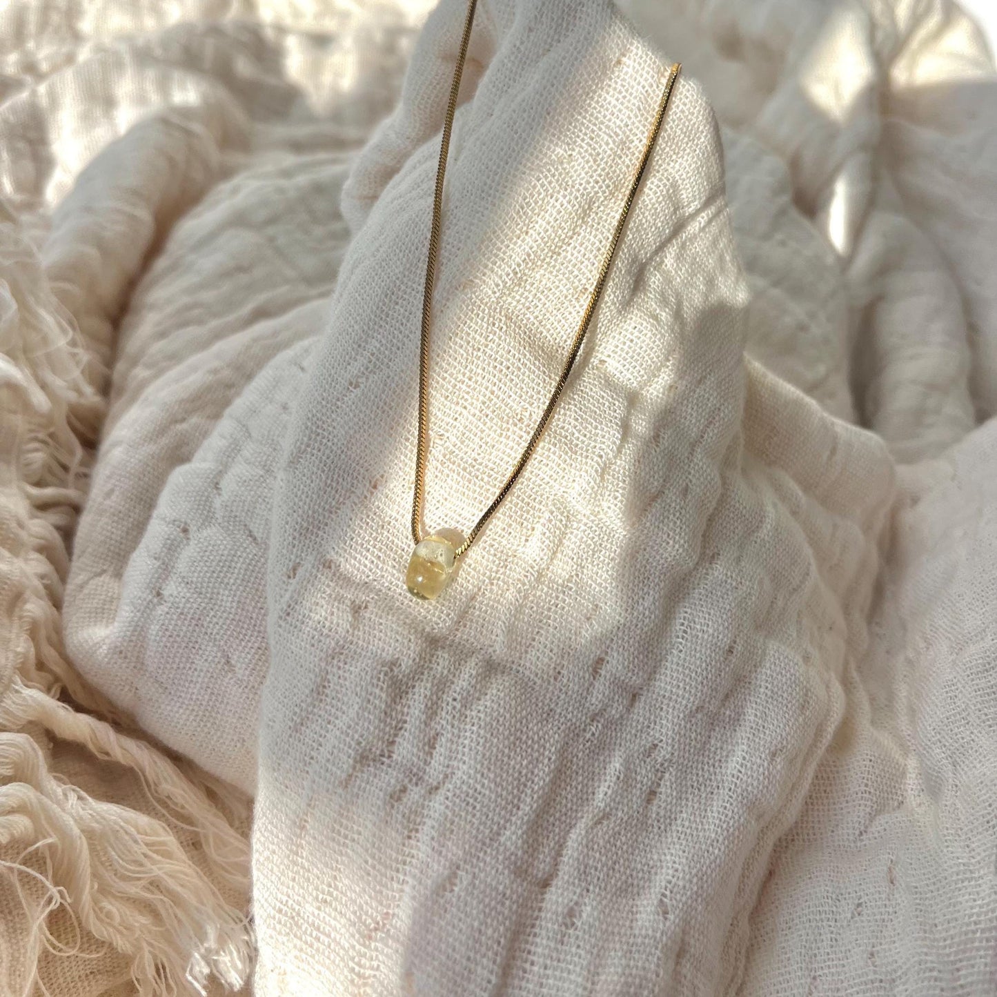 
                  
                    Delicate gold necklace with a small yellow gemstone on a textured beige fabric background
                  
                