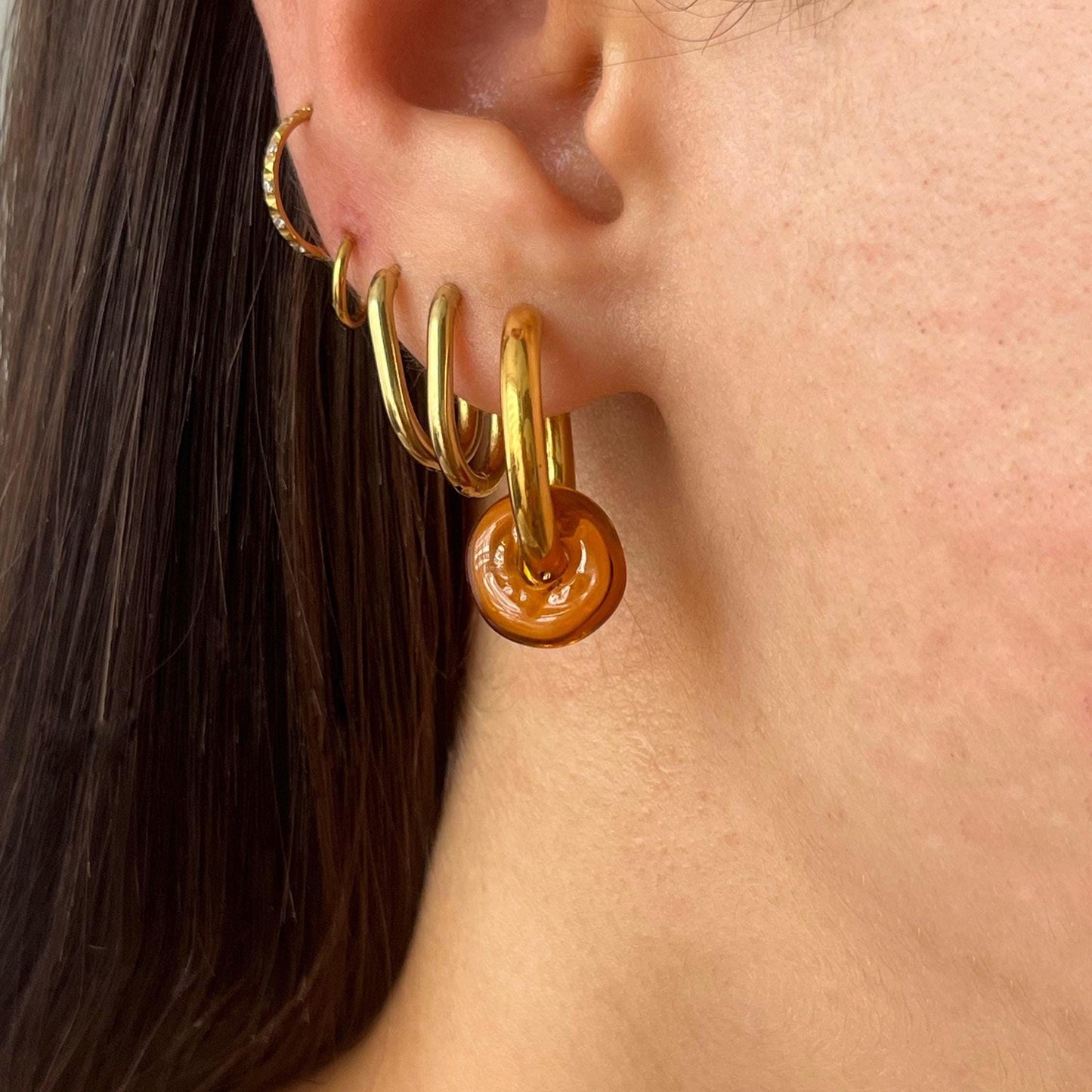 
                  
                    Close-up of an ear wearing gold hoop earrings with a brown circular earring.
                  
                