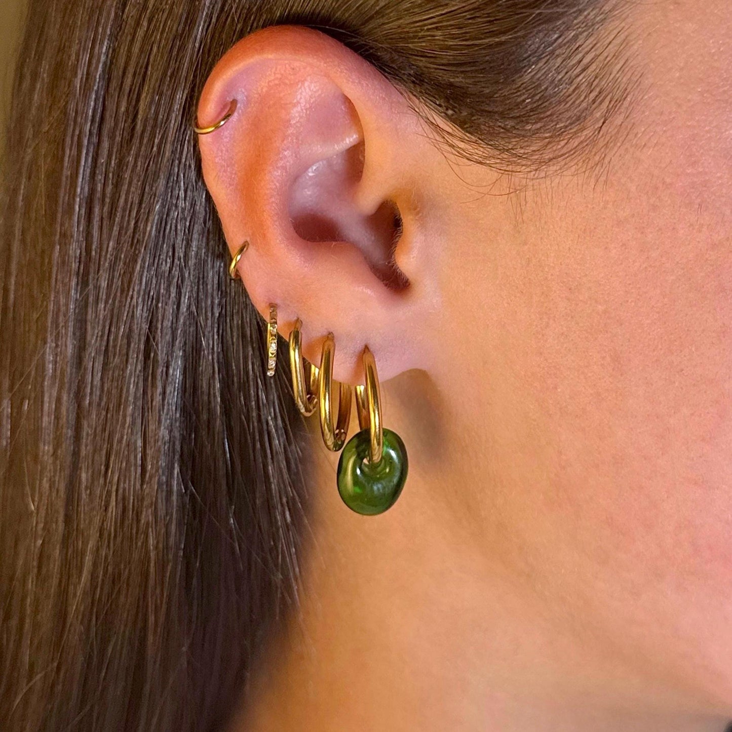 
                  
                    Gold hoop earrings with a green bead on an ear against a blurred background
                  
                