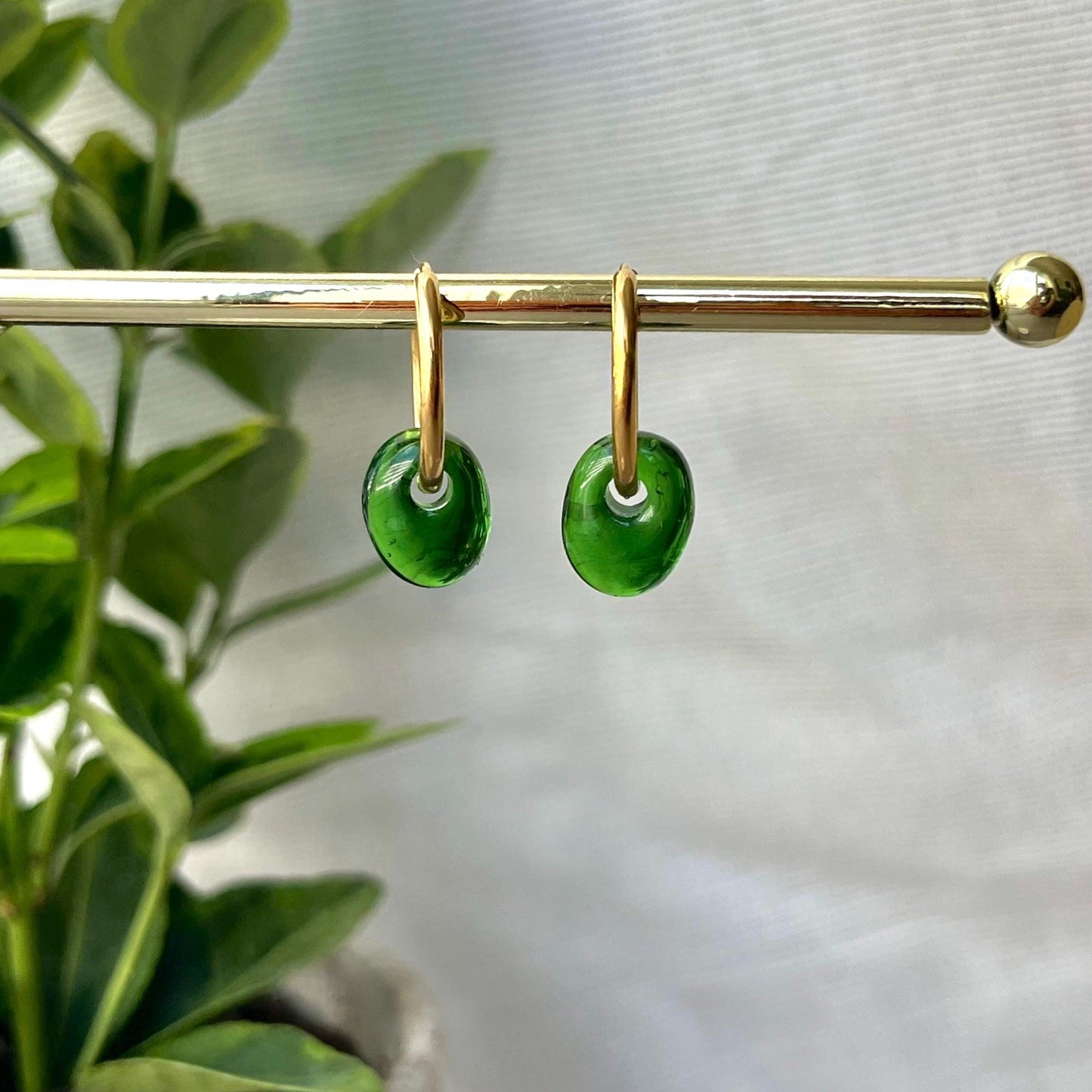 
                  
                    Green earrings on a gold hoop against a blurred green plant background
                  
                