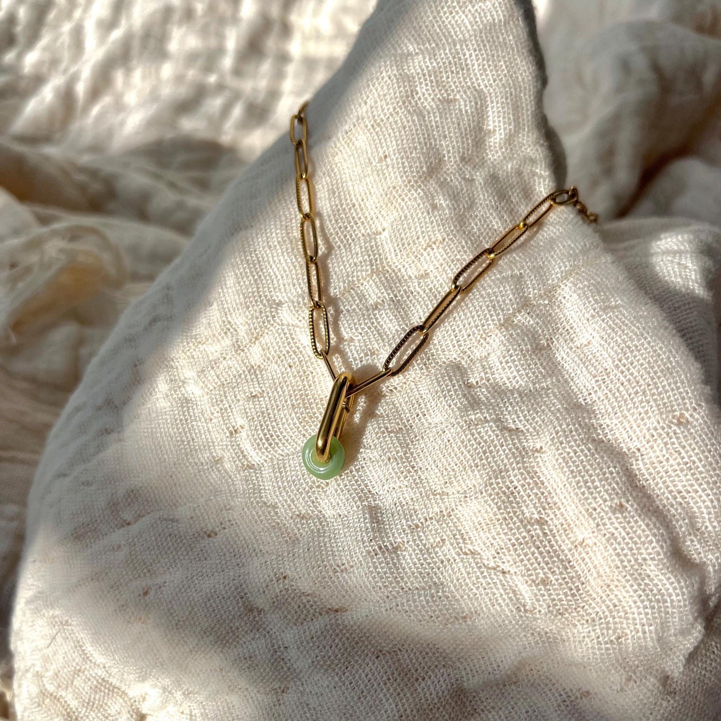 
                  
                    Gold chain necklace with a green pendant on a textured fabric background
                  
                