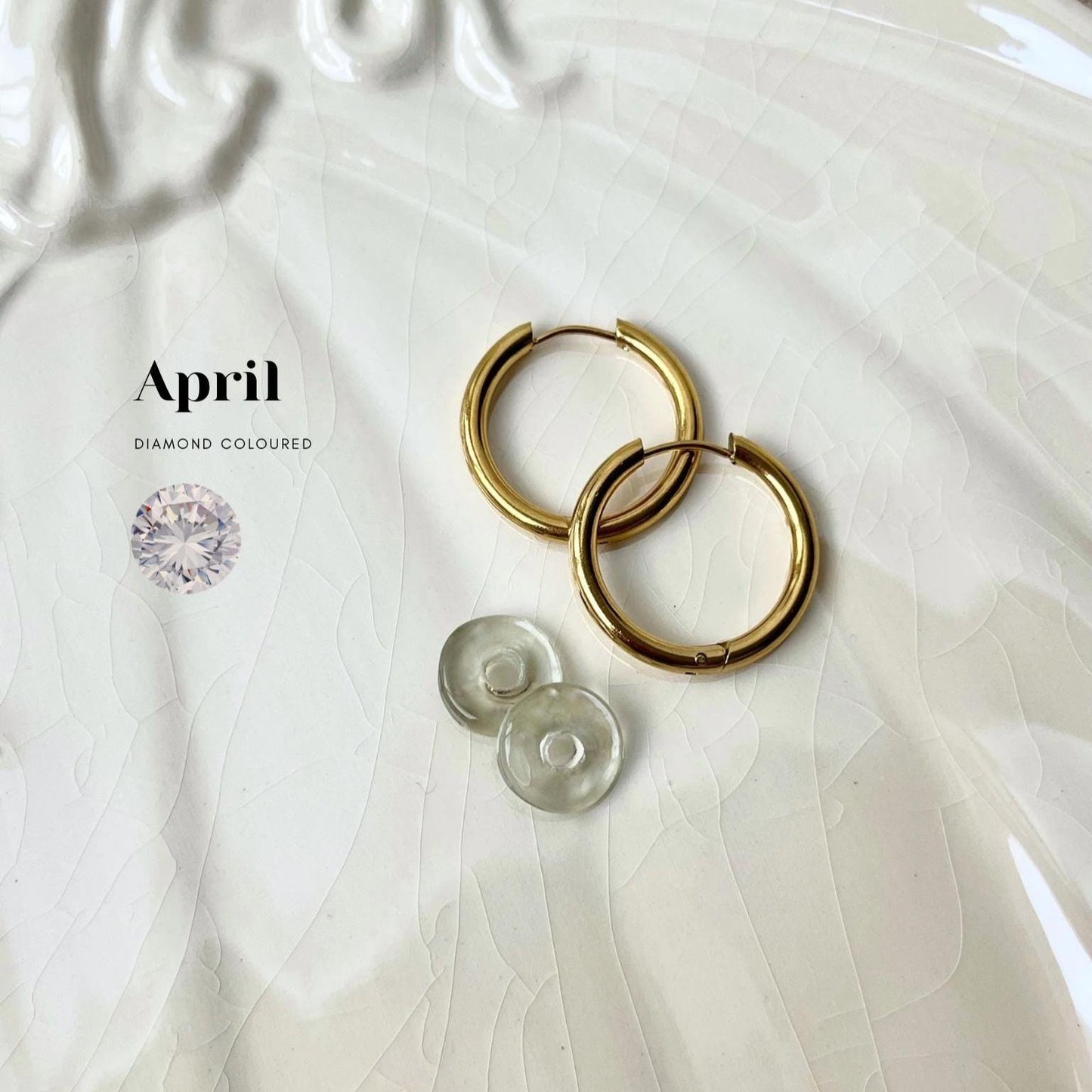 
                  
                    Gold hoop earrings on a textured white surface with 'April' and 'Diamond Coloured' text.
                  
                