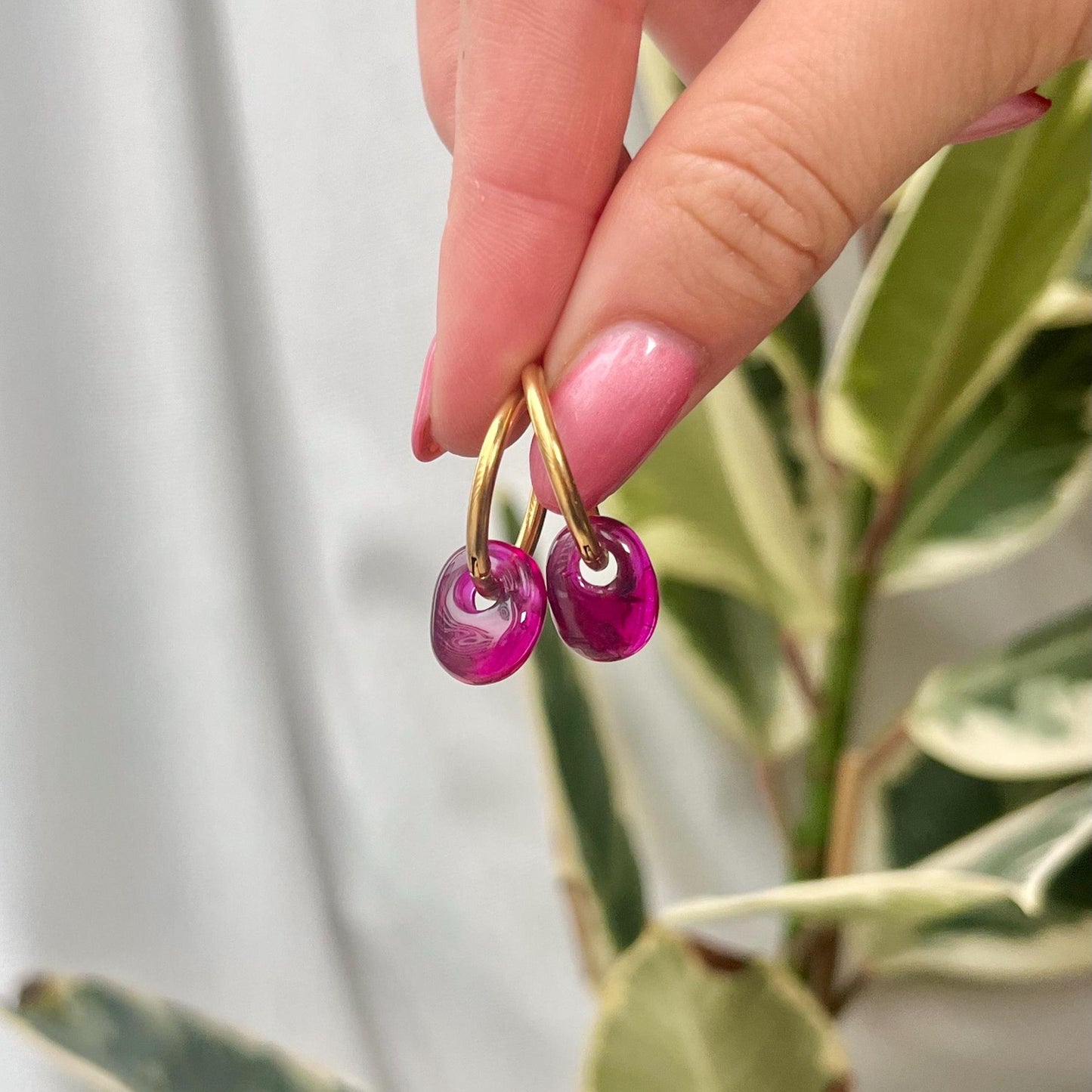 Hand holding a pair of gold earrings with pink gemstones against a blurred green plant background