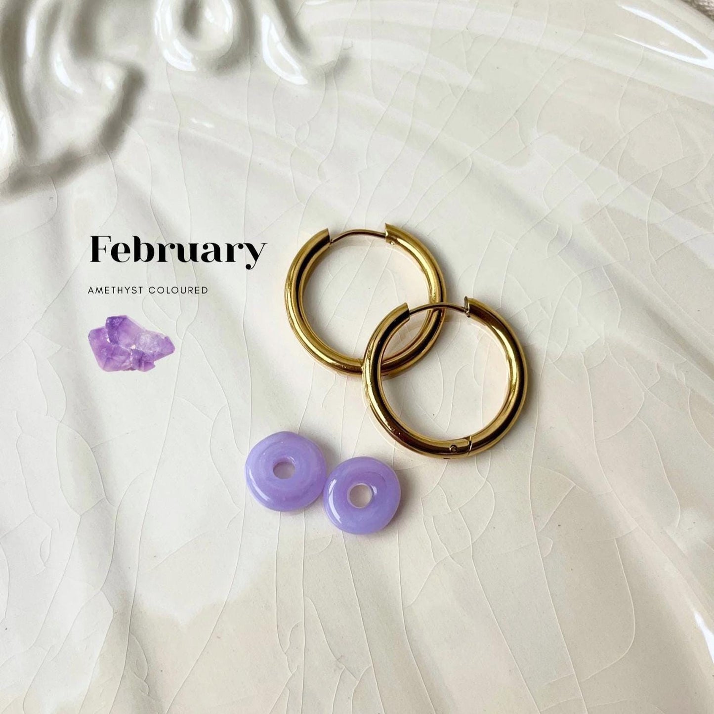
                  
                    Gold hoop earrings with purple gemstones on a textured white surface, featuring the word 'February'.
                  
                