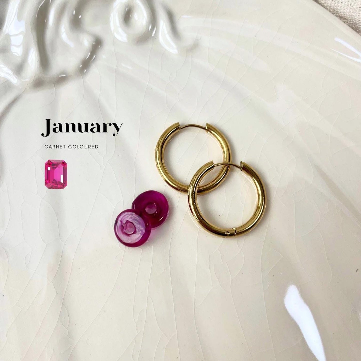 Gold hoop earrings with garnet-colored gemstones on a textured white surface, featuring the word 'January'.