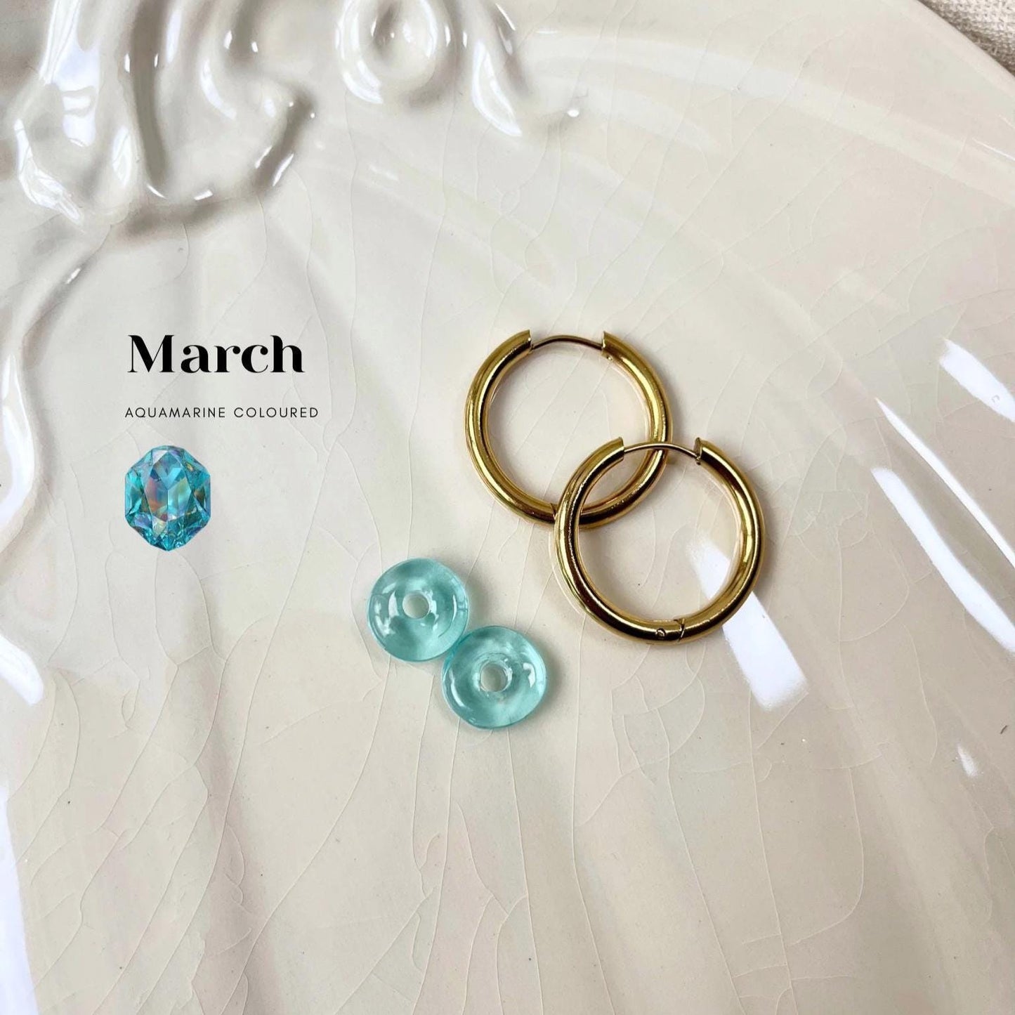 
                  
                    Gold hoop earrings with aquamarine-colored stones on a textured white surface, with 'March' and 'Aquamarine Coloured' text.
                  
                